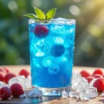 A tall glass of Blue Razz Ice Juice garnished with mint and raspberries on a wooden table with sunlight streaming in.