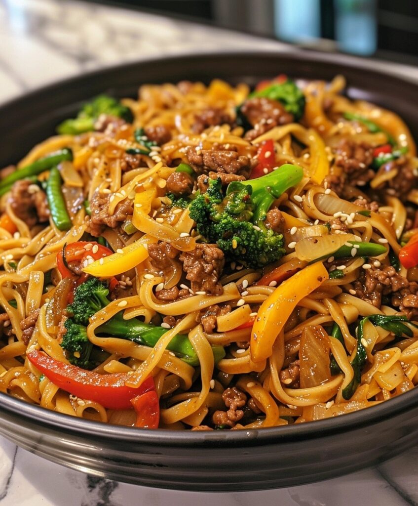 A bowl of Sticky Beef Noodles with broad noodles, ground beef, broccoli florets, bell peppers, green beans, and sesame seeds, showcasing a colorful and hearty Asian dish.


