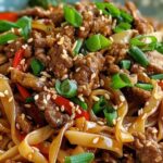 A plate of Sticky Beef Noodles with broad noodles, tender beef strips, colorful vegetables, sesame seeds, and green onions, showcasing a flavorful Asian dish.